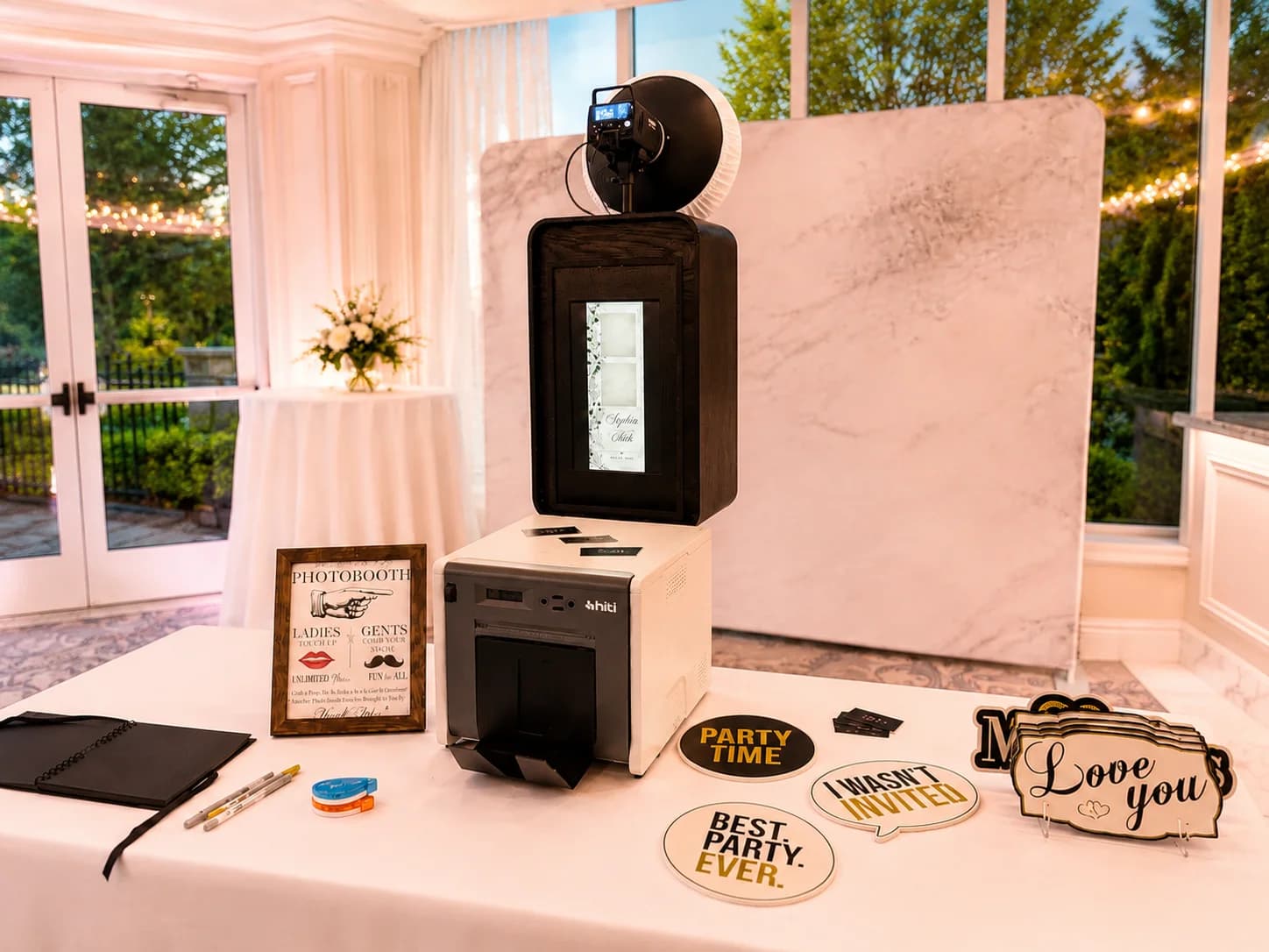 A DSLR photo booth, professional printer, guestbook, and props arranged in a bright garden room