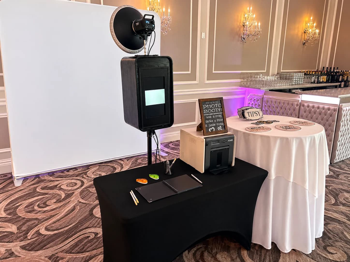 A black DSLR photo booth with printer, guestbook, props, and white backdrop in an elegant ballroom