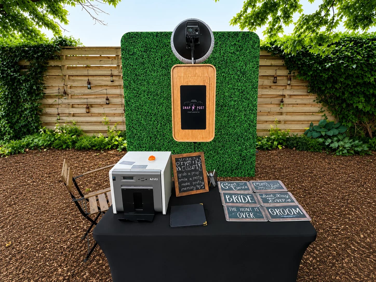 A wood-finish photo booth, printer, guestbook, props, and green hedge backdrop at an outdoor event