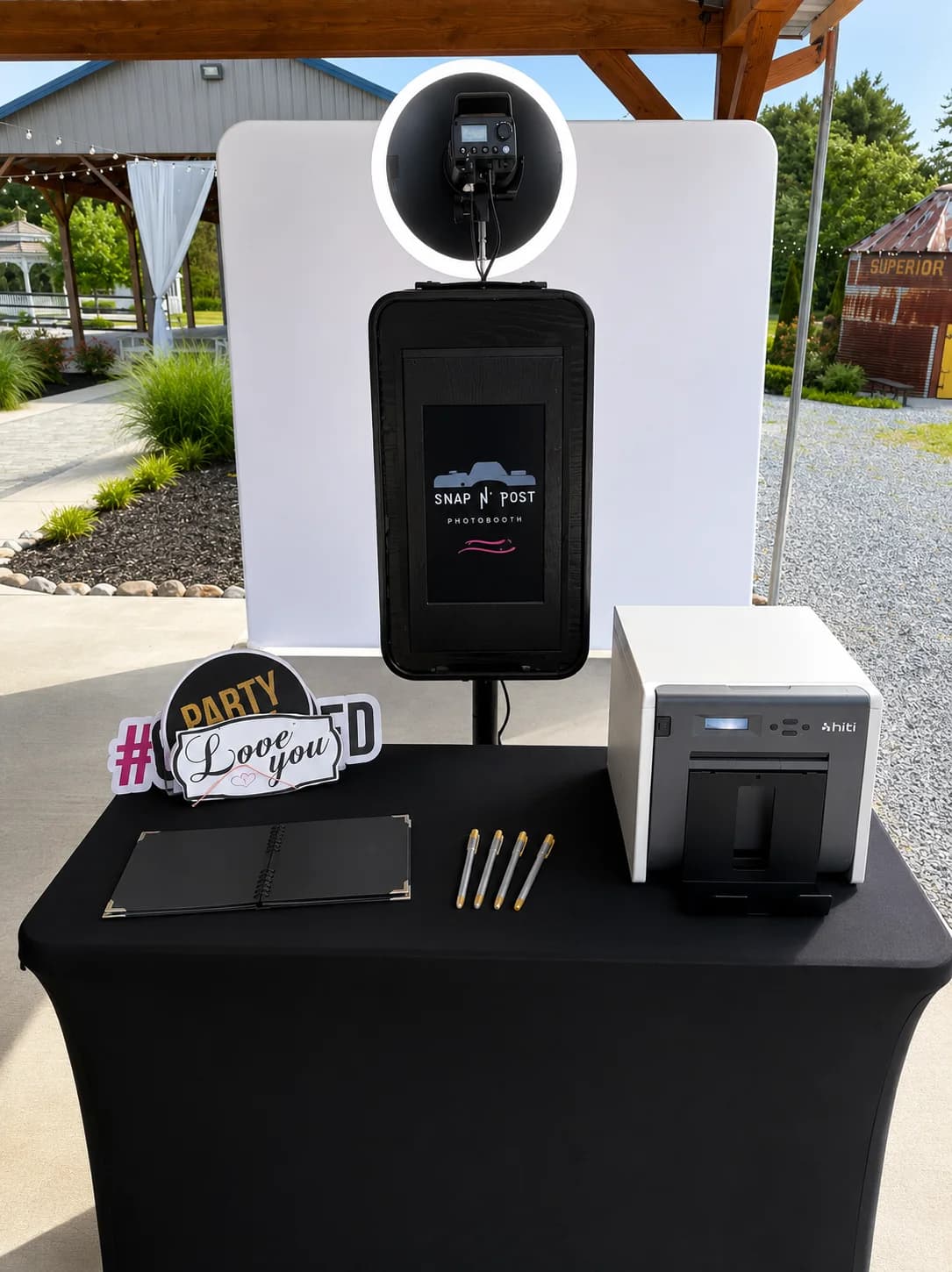 A DSLR photo booth, printer, guestbook, and props arranged under an outdoor pavilion